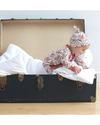 Baby in a patterned outfit and hat lying in an open suitcase on a light wooden floor.