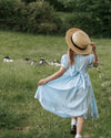Person in a blue dress and straw hat walking through a field with animals.