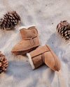 Pair of brown children's boots with white fur lining on a textured surface with pine cones.