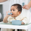 Baby sitting in a high chair with blue utensils and bowl, being fed by an adult.