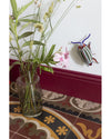 Clear vase with greenery on a patterned floor next to a white wall with a decorative beetle.