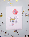 A white greeting card featuring a whimsical illustration of a white horse upside down holding a red balloon with a blue number 6, displayed on a light gray background decorated with scattered gold and silver star confetti.