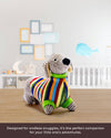 Colorful stuffed dog toy on a wooden floor with a baby crib and toys in the background.