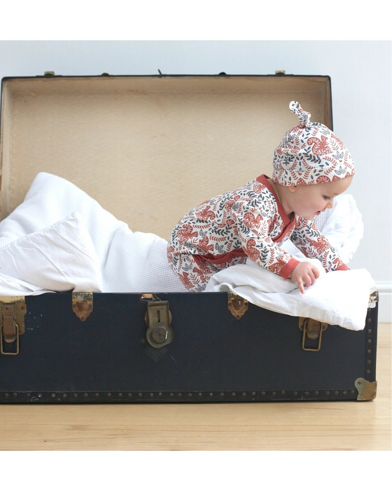 Baby in a patterned outfit and hat lying in an open suitcase on a light wooden floor.
