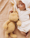 Baby in a white hooded outfit holding a plush bunny toy on a beige blanket