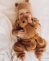 Baby in a brown bear onesie holding a teddy bear on a soft white background