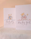 Two white baby shower cards standing upright on a white fabric surface against a warm beige wall. The left card says 'baby boy' with a teddy bear illustration in blue.
