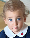 A close-up portrait of a toddler with light brown hair and bright blue eyes, wearing a navy blue top with a white Peter Pan collar featuring a small red apple embroidery.