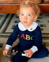 A toddler sitting on a patterned rug indoors, wearing a navy blue footed onesie with a white Peter Pan collar, colorful embroidered letters 'ABC' on the chest, and a small red apple detail.