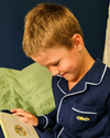 A young boy with short blond hair wearing navy blue pajamas with white piping and a small yellow car embroidery on the chest pocket, looking down at a book in his hands.