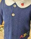 Close-up of a navy blue ribbed knit baby top with a white collar featuring a red apple embroidery, two silver buttons, and colorful embroidered letters 'ABC' on the chest.