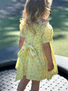 A young girl viewed from behind wearing a yellow short-sleeve dress with a small floral print and a tie waist, standing outdoors on a paved surface with grass in the background.