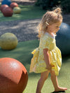 A young toddler girl with blonde hair in a small bun wearing a yellow patterned dress, walking barefoot on green artificial turf in an outdoor playground setting with large decorative spheres.