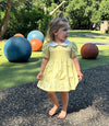 A toddler girl wearing a light yellow smocked dress with a white Peter Pan collar stands barefoot on an outdoor playground path. Large colorful exercise balls and green grass are visible in the background.