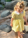 A young girl with blonde hair in a messy bun stands barefoot on a rocky outdoor path, wearing a yellow short-sleeve dress with a white ruffled collar.