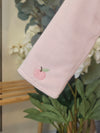 A close-up of a light pink ribbed fabric sleeve with a small embroidered pink apple and green leaf detail, set against a soft-focus background of green leaves and white flowers.