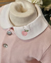 A close-up of a baby clothing display showing a light pink ribbed knit top with a wide cream-colored collar. The collar is embroidered with a small pink apple and green leaf, and has silver buttons.