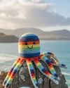 Colorful crochet octopus toy on a rock with a scenic background of water and mountains.