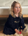 A young girl with blonde hair sits on a beige rug in front of a cream-colored sofa, wearing a black long-sleeve dress with floral embroidery on the chest.
