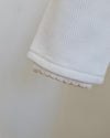 Close-up of a white ribbed knit fabric cuff with a delicate beige scalloped trim along the edge, set against a plain light beige background.