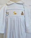 A white baby smocked dress with short sleeves, a Peter Pan collar, and a smocked chest panel featuring embroidered toys including a rocking horse, a yellow duck, and a teddy bear.