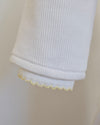 Close-up of a white ribbed knit sleeve or cuff with a delicate yellow scalloped trim along the inner edge, shown against a soft light beige background.