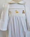 White baby smocked dress with a matching white cardigan, featuring a smocked chest panel with embroidered yellow duck, brown teddy bear, and gold rocking horse motifs.