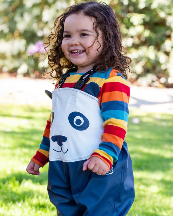 Child wearing a colorful striped shirt and blue overalls with a white bag featuring a dog face design, standing outdoors.