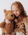 Child holding a large brown teddy bear with a smaller teddy bear attached to their shirt against a white background