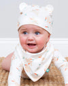 Baby wearing a white hat with bear ears and a matching bib with animal prints on a neutral background.