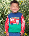 Child wearing a colorful sweater with a fire truck design, standing in front of green foliage.