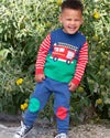 Child wearing a colorful outfit with a fire truck design in front of green foliage.