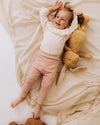 Child lying on a bed with teddy bears, wearing a white sweater and pink pants.