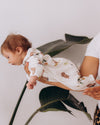 Baby in a floral outfit held by an adult against a white background with a plant.