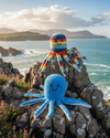 Two crocheted octopus toys on a rocky outcrop with ocean and mountains in the background