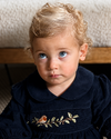 A toddler with curly blonde hair and blue eyes wearing a dark navy blue velvet dress with a Peter Pan collar and embroidered gold leaf and bird motif on the chest, posed against a neutral indoor background.
