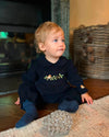 A toddler sitting on a wooden floor in a cozy room, wearing a navy blue long-sleeve romper with a floral embroidered chest detail and matching navy socks.