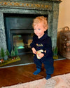 A toddler with curly blonde hair wearing a navy blue embroidered jumpsuit and blue socks stands in front of a fireplace, holding a small yellow-green toy.