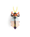 A colorful beetle-shaped wall sculpture or decorative object shown from above on a plain white background. The design features a striped cream and orange wing case, a dark central body, yellow horns, and orange and blue accents.