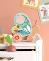 Colorful butterfly-shaped wooden toy on a shelf with a pink wall and framed pictures in the background.