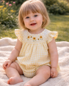 Child wearing a yellow checkered outfit with sunflower details, sitting on a blanket outdoors.