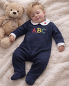 Child wearing a navy blue outfit with colorful letters, lying next to a teddy bear on a soft surface.