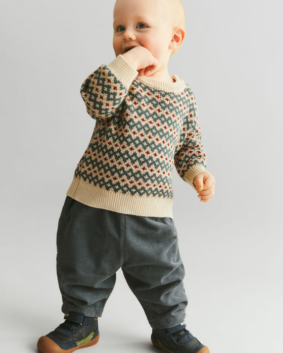 Baby wearing a patterned sweater and dark pants on a plain background