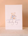 A vertical baby shower card featuring a watercolor illustration of a teddy bear wearing a blue bow, above the cursive text 'baby boy' in dark blue ink, set against a warm beige background.