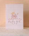 A white baby shower card featuring a watercolor illustration of a teddy bear wearing a gold crown and holding a pink bow, with the words 'baby girl' written in elegant cursive script below, set against a warm beige background.