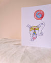 A white greeting card standing upright on a light fabric surface against a warm beige background. The card features a whimsical watercolor illustration of a white pig wearing a yellow and blue party hat and holding a red balloon with a blue spiral design.