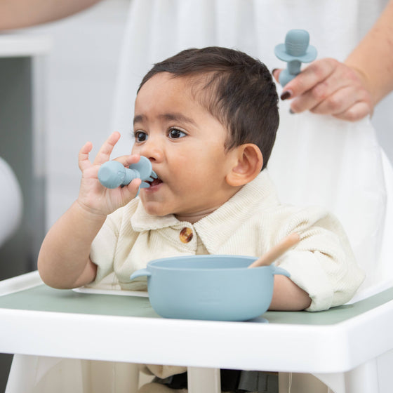 Baby sitting in a high chair with blue utensils and bowl, being fed by an adult.