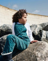 Child sitting on rocks wearing teal overalls and white shirt, with a brick wall and blue sky in the background.