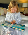 A young girl with blonde hair sits on a bed, reading a colorful picture book. She is wearing a white long-sleeve nightgown with embroidered details on the chest.